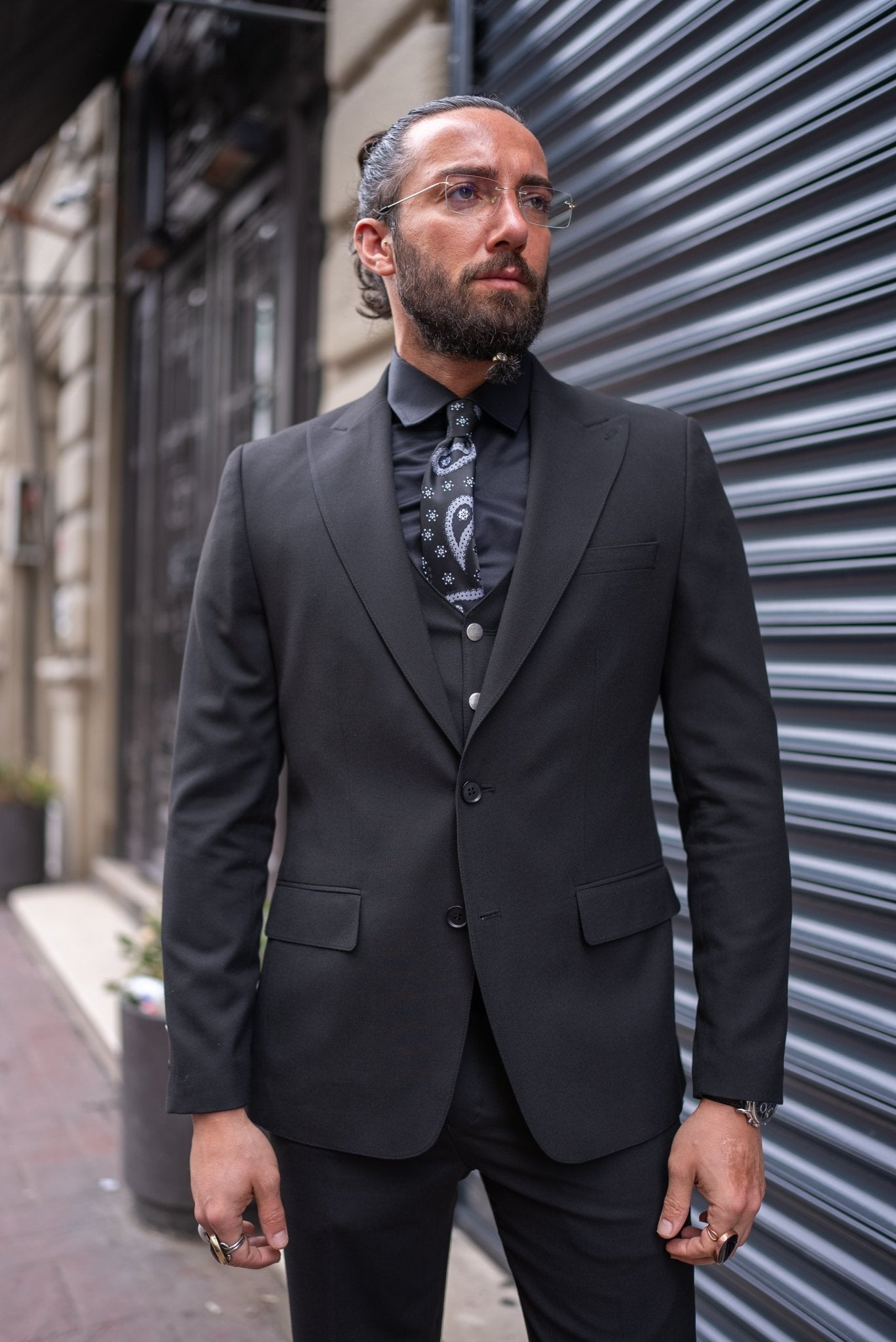 A man is confidently dressed in a black slim-fit tailored suit, standing against an urban backdrop with a metallic shutter. He pairs the suit with a black shirt, black tie with subtle patterns, and a matching black vest. His look is completed with polished black dress shoes and a black ring on one hand. He has a well-groomed beard, clear glasses, and his hair is tied back. The man's pose, with one leg slightly raised, exudes sophistication and a sharp, modern style.