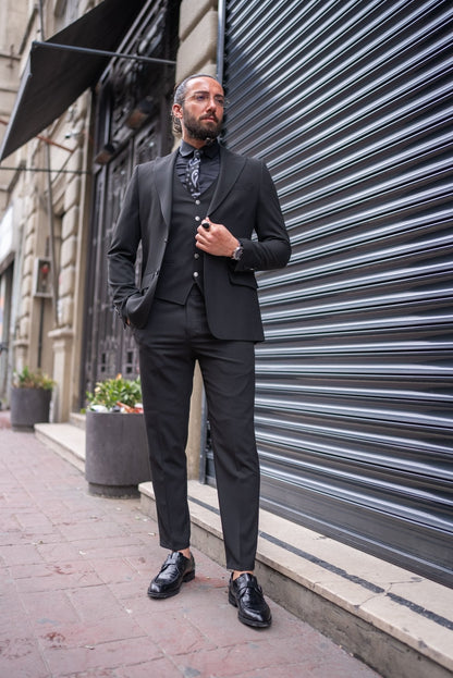 A man is confidently dressed in a black slim-fit tailored suit, standing against an urban backdrop with a metallic shutter. He pairs the suit with a black shirt, black tie with subtle patterns, and a matching black vest. His look is completed with polished black dress shoes and a black ring on one hand. He has a well-groomed beard, clear glasses, and his hair is tied back. The man's pose, with one leg slightly raised, exudes sophistication and a sharp, modern style.