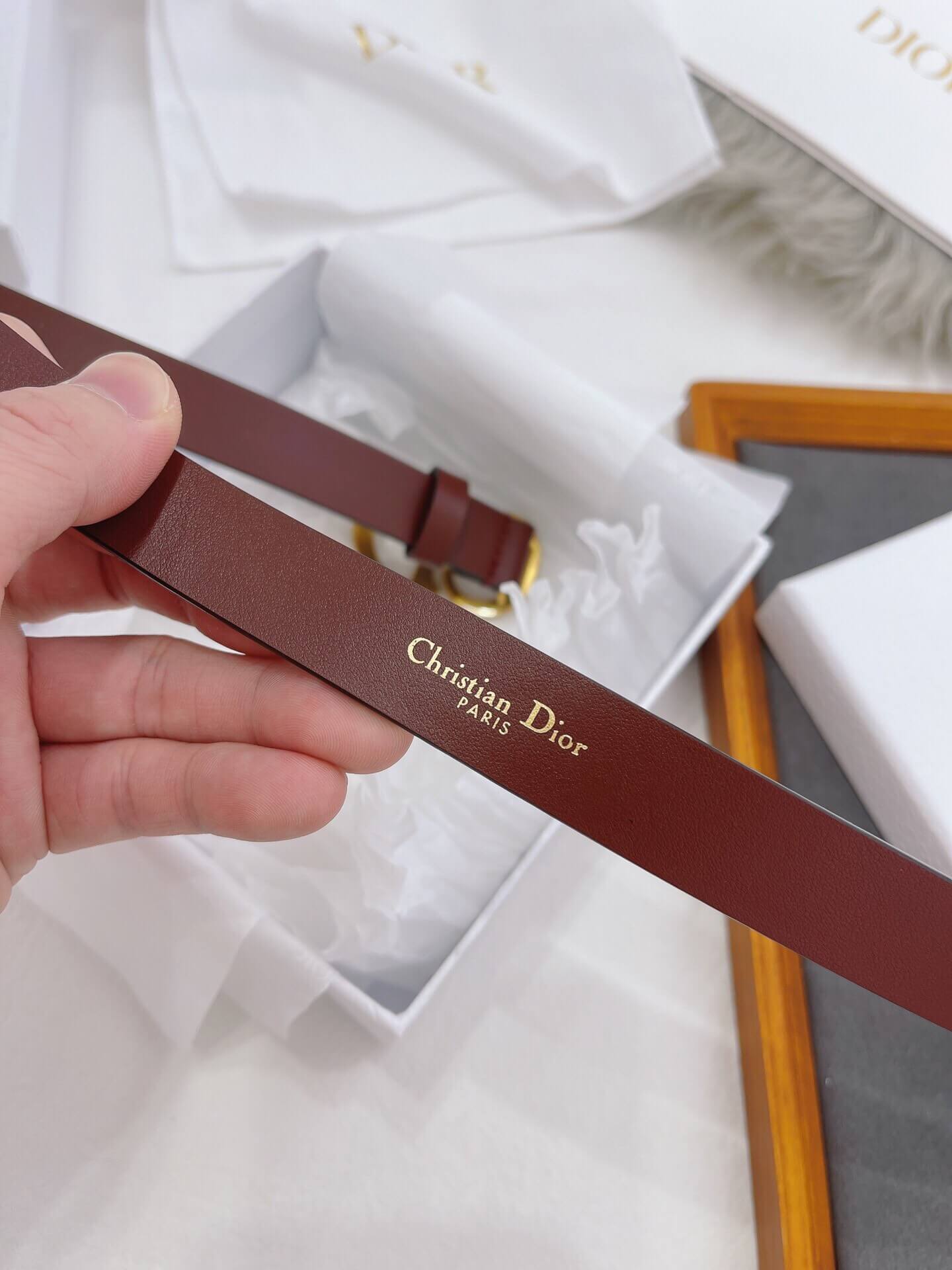 Brown leather belt with 'Christian Dior' logo held by a hand, with a white background.