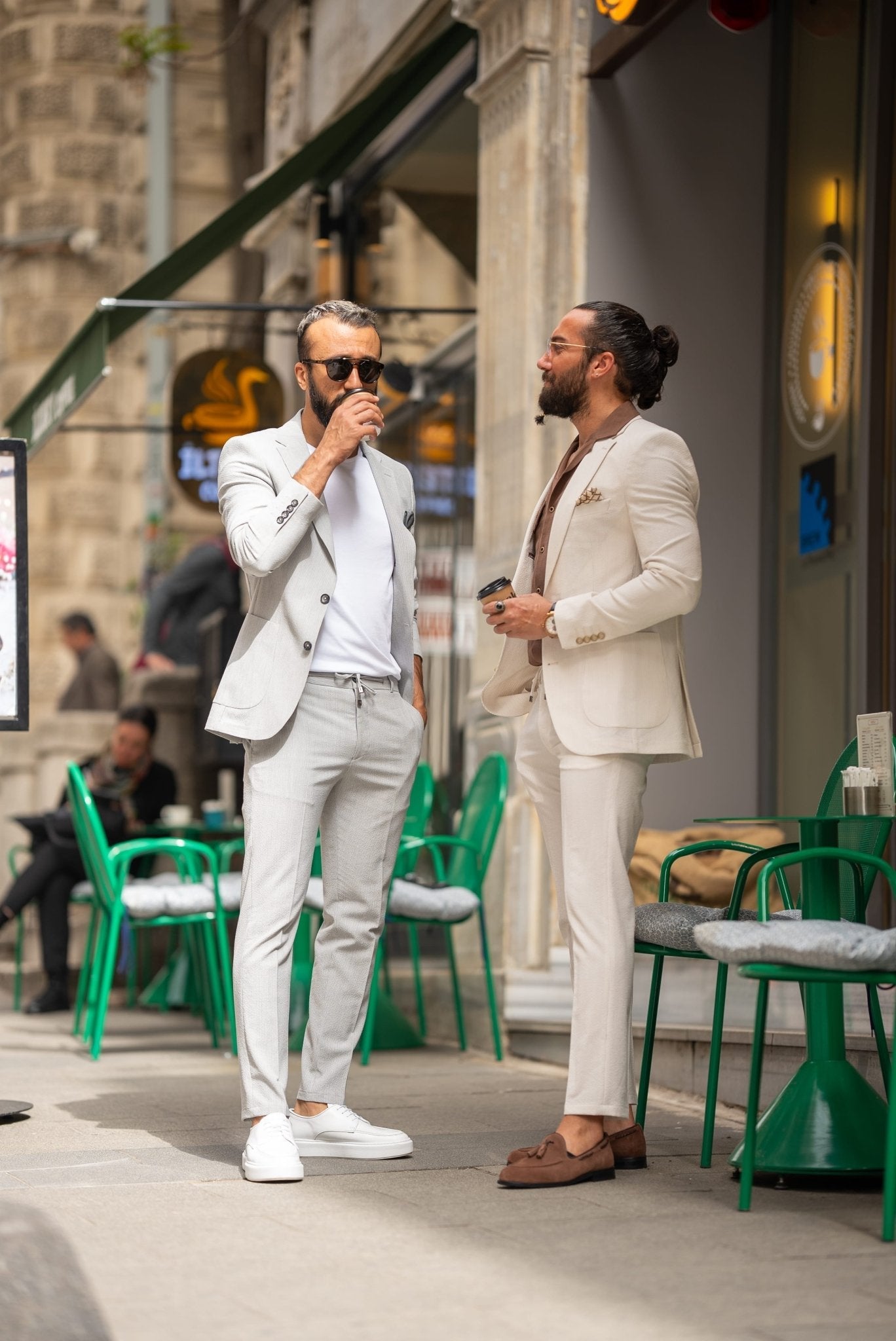 Two men are casually dressed in light-colored suits, standing on an outdoor café terrace. The man on the left wears a light grey seersucker suit with a white t-shirt and white sneakers, holding a coffee cup in one hand. The man on the right is in a beige suit with a brown shirt underneath and brown loafers, leaning on a green café chair. Both men have beards and are sporting sunglasses, exuding a stylish yet relaxed vibe. The scene is set against an urban backdrop with green café chairs around them.