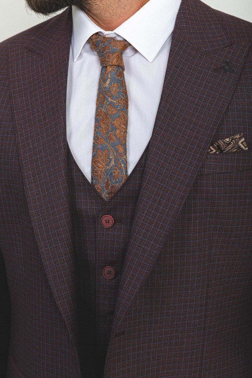 A well-groomed man wearing a Burgundy Checkered Three-Piece Suit stands confidently in a studio setting. The suit features a rich burgundy color with a subtle checkered pattern, paired with a white dress shirt, a patterned tie, and a matching pocket square, adding a refined touch to the ensemble. Brown leather dress shoes complement the outfit, enhancing its classic elegance. The man has neatly styled hair and a trimmed beard, projecting a sophisticated and polished look ideal for formal occasion. 