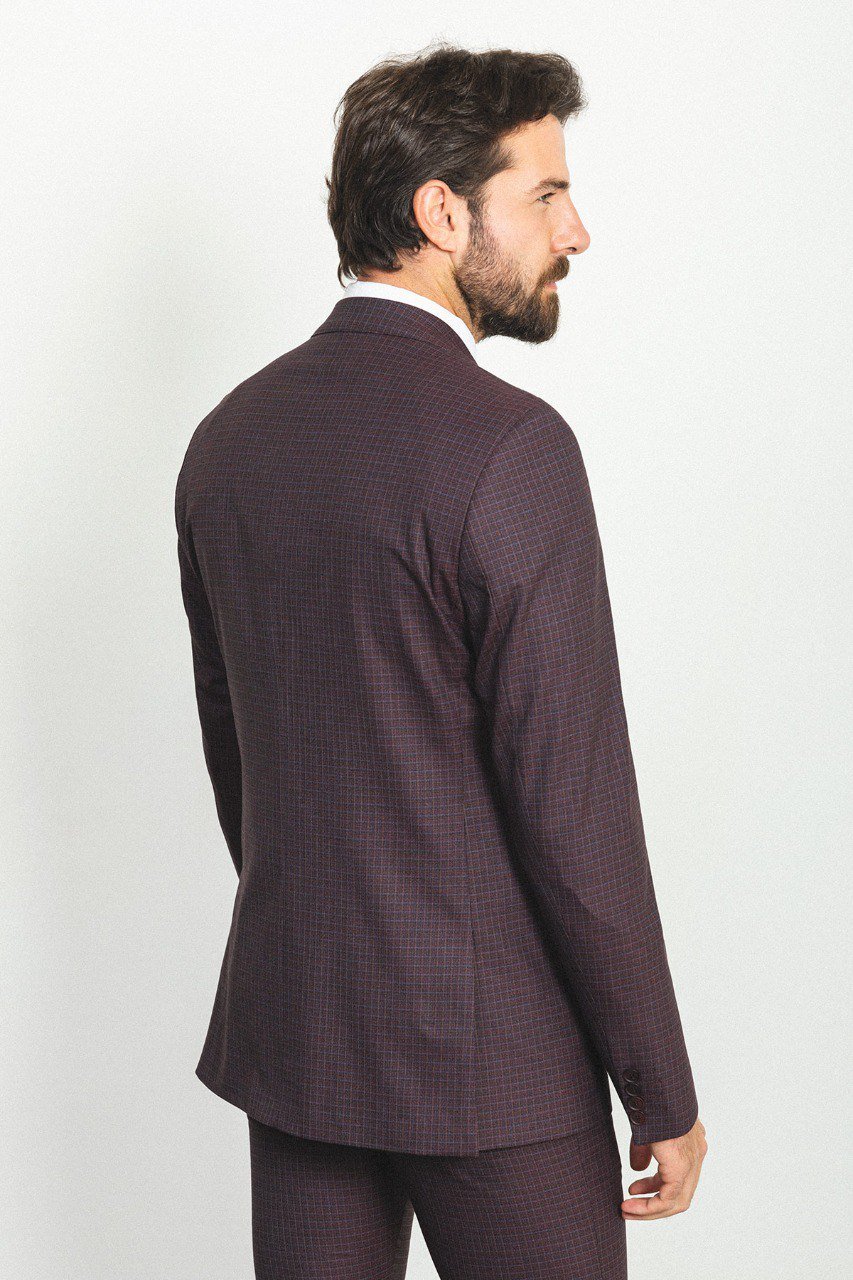 A well-groomed man wearing a Burgundy Checkered Three-Piece Suit stands confidently in a studio setting. The suit features a rich burgundy color with a subtle checkered pattern, paired with a white dress shirt, a patterned tie, and a matching pocket square, adding a refined touch to the ensemble. Brown leather dress shoes complement the outfit, enhancing its classic elegance. The man has neatly styled hair and a trimmed beard, projecting a sophisticated and polished look ideal for formal occasions.