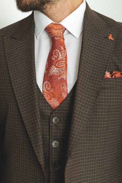 A well-dressed man wearing a Brown Checkered Three-Piece Suit stands confidently in a studio setting. The suit features a subtle checkered pattern, paired with a white dress shirt, a vibrant red paisley tie, and a matching pocket square, adding a touch of sophistication. The ensemble is completed with brown leather dress shoes, exuding classic elegance. The man has neatly groomed hair and a trimmed beard, embodying a polished and refined look perfect for formal occasions.