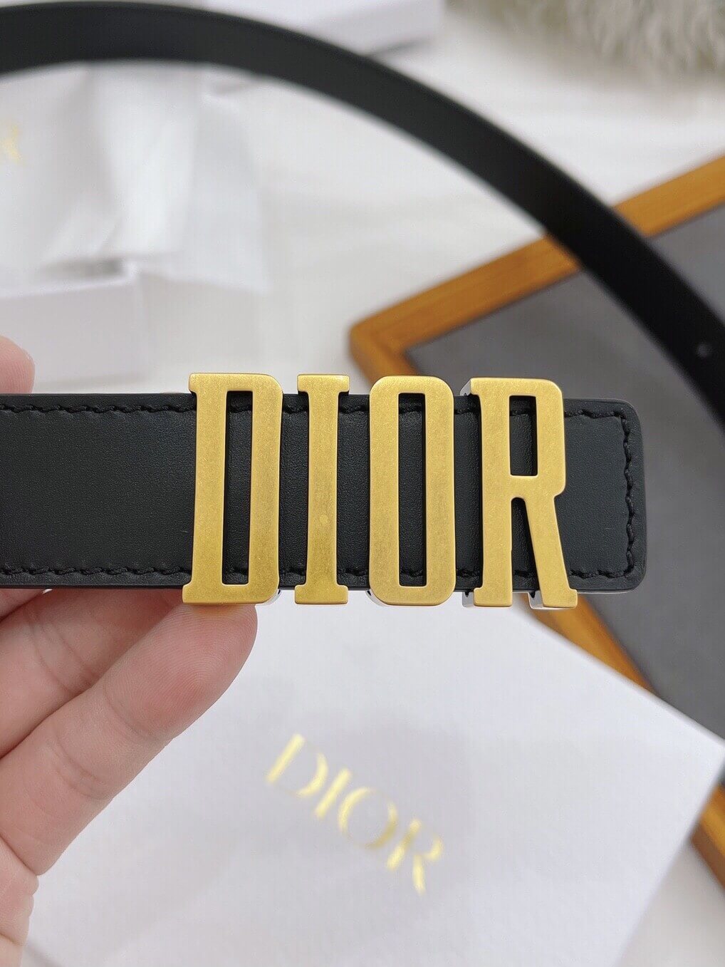 Black Dior belt with gold logo held by a hand, with a white Dior box in the background.