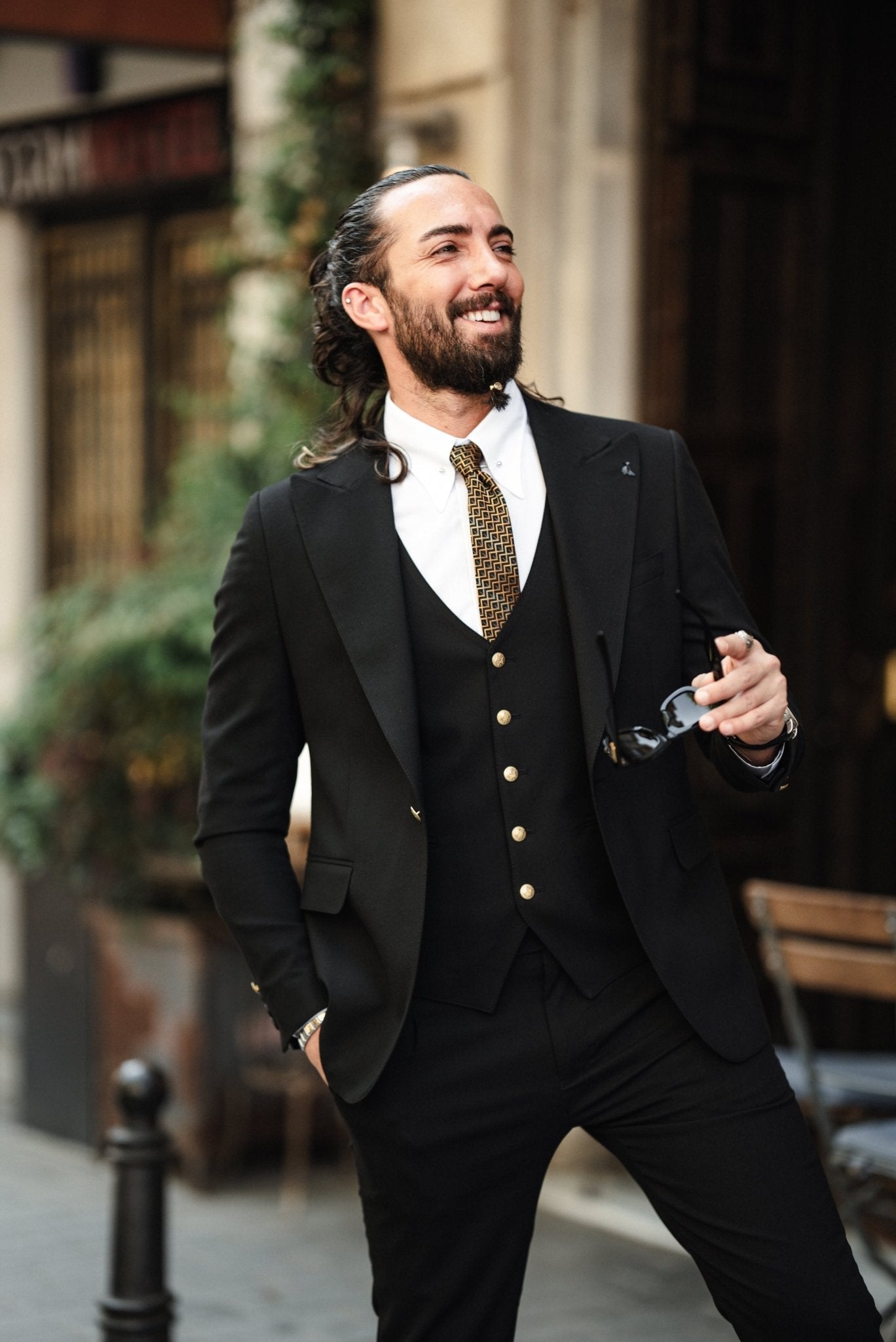 A fashionable man wearing an Acadia Black Slim-Fit Suit exudes confidence on a city street. The suit is impeccably tailored, featuring gold buttons on the vest and jacket, paired with a crisp white dress shirt and a patterned tie. The man accessorizes with stylish sunglasses, a statement ring, and a luxury watch, enhancing his sophisticated look. His neatly groomed beard and pulled-back hair add to his sharp and modern appearance.