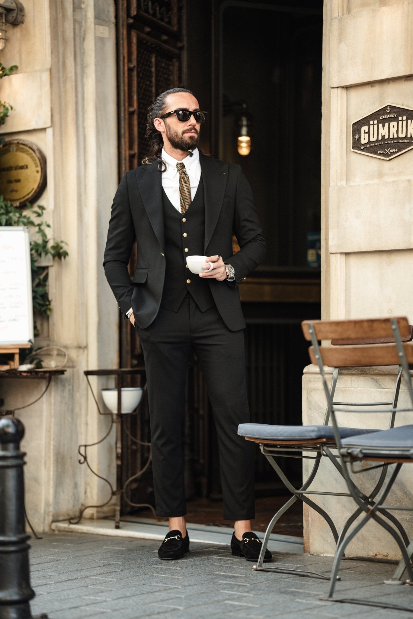 A fashionable man wearing an Acadia Black Slim-Fit Suit exudes confidence on a city street. The suit is impeccably tailored, featuring gold buttons on the vest and jacket, paired with a crisp white dress shirt and a patterned tie. The man accessorizes with stylish sunglasses, a statement ring, and a luxury watch, enhancing his sophisticated look. His neatly groomed beard and pulled-back hair add to his sharp and modern appearance.