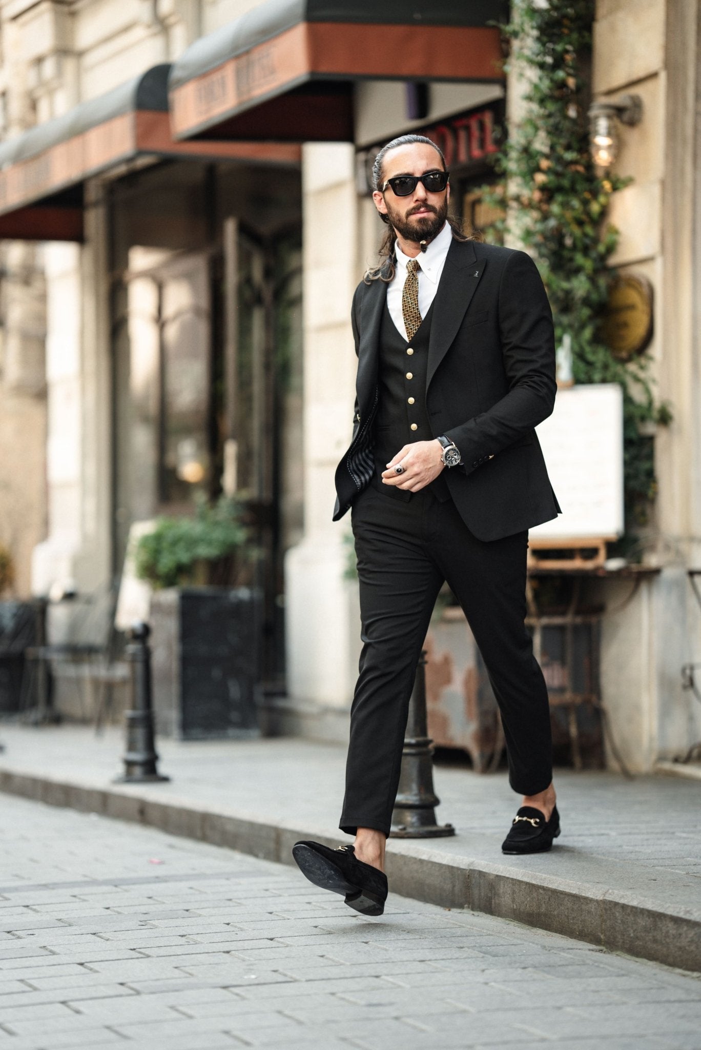 A fashionable man wearing an Acadia Black Slim-Fit Suit exudes confidence on a city street. The suit is impeccably tailored, featuring gold buttons on the vest and jacket, paired with a crisp white dress shirt and a patterned tie. The man accessorizes with stylish sunglasses, a statement ring, and a luxury watch, enhancing his sophisticated look. His neatly groomed beard and pulled-back hair add to his sharp and modern appearance.