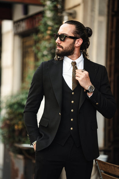 A fashionable man wearing an Acadia Black Slim-Fit Suit exudes confidence on a city street. The suit is impeccably tailored, featuring gold buttons on the vest and jacket, paired with a crisp white dress shirt and a patterned tie. The man accessorizes with stylish sunglasses, a statement ring, and a luxury watch, enhancing his sophisticated look. His neatly groomed beard and pulled-back hair add to his sharp and modern appearance.