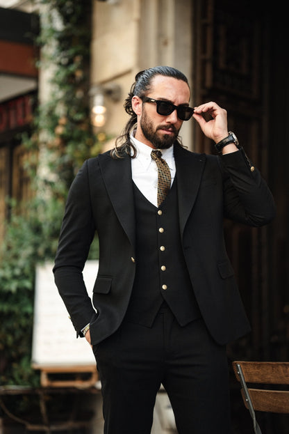 A fashionable man wearing an Acadia Black Slim-Fit Suit exudes confidence on a city street. The suit is impeccably tailored, featuring gold buttons on the vest and jacket, paired with a crisp white dress shirt and a patterned tie. The man accessorizes with stylish sunglasses, a statement ring, and a luxury watch, enhancing his sophisticated look. His neatly groomed beard and pulled-back hair add to his sharp and modern appearance.