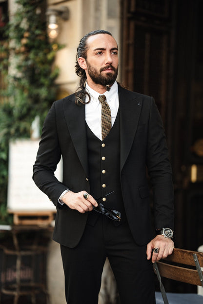 A fashionable man wearing an Acadia Black Slim-Fit Suit exudes confidence on a city street. The suit is impeccably tailored, featuring gold buttons on the vest and jacket, paired with a crisp white dress shirt and a patterned tie. The man accessorizes with stylish sunglasses, a statement ring, and a luxury watch, enhancing his sophisticated look. His neatly groomed beard and pulled-back hair add to his sharp and modern appearance.