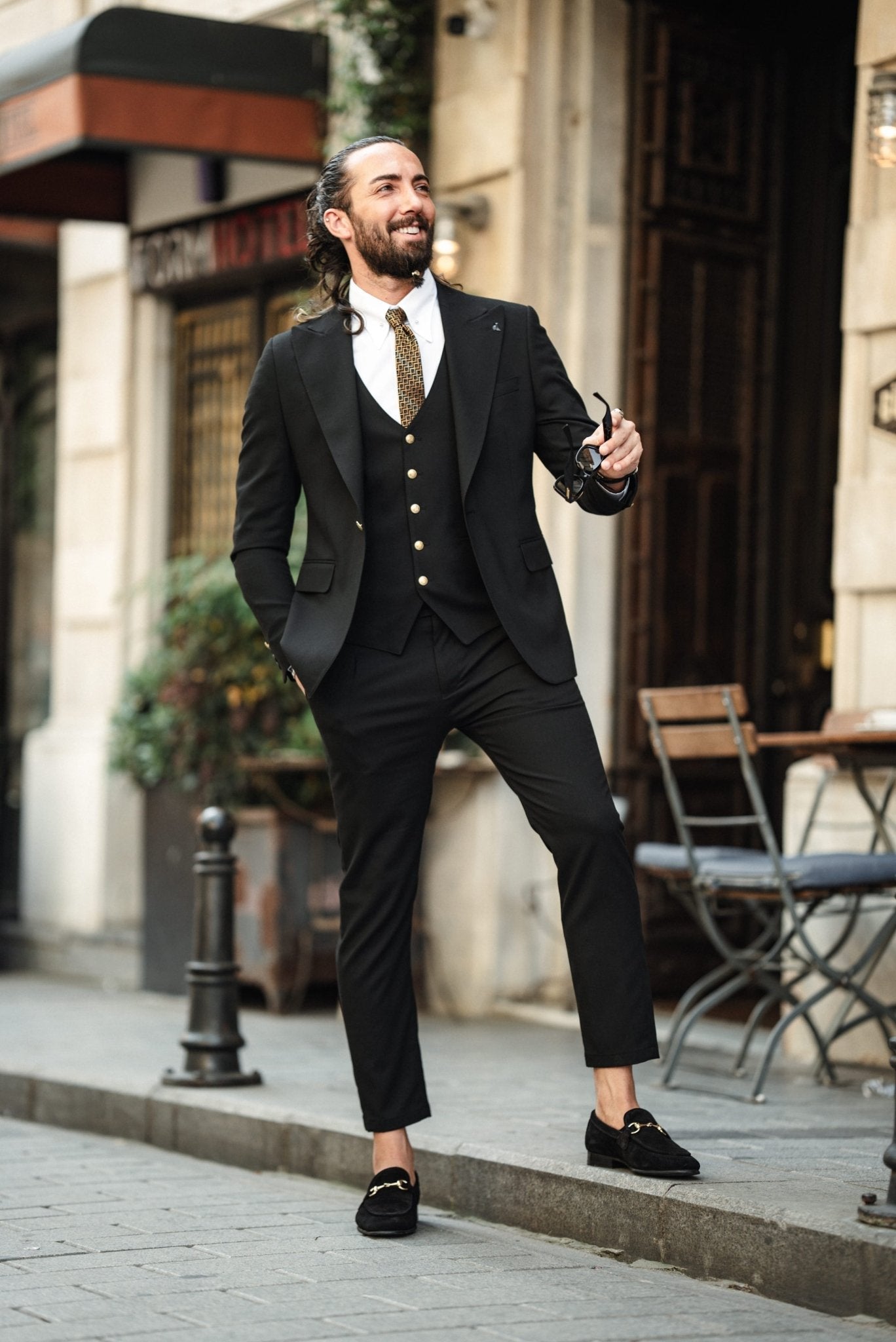 A fashionable man wearing an Acadia Black Slim-Fit Suit exudes confidence on a city street. The suit is impeccably tailored, featuring gold buttons on the vest and jacket, paired with a crisp white dress shirt and a patterned tie. The man accessorizes with stylish sunglasses, a statement ring, and a luxury watch, enhancing his sophisticated look. His neatly groomed beard and pulled-back hair add to his sharp and modern appearance.