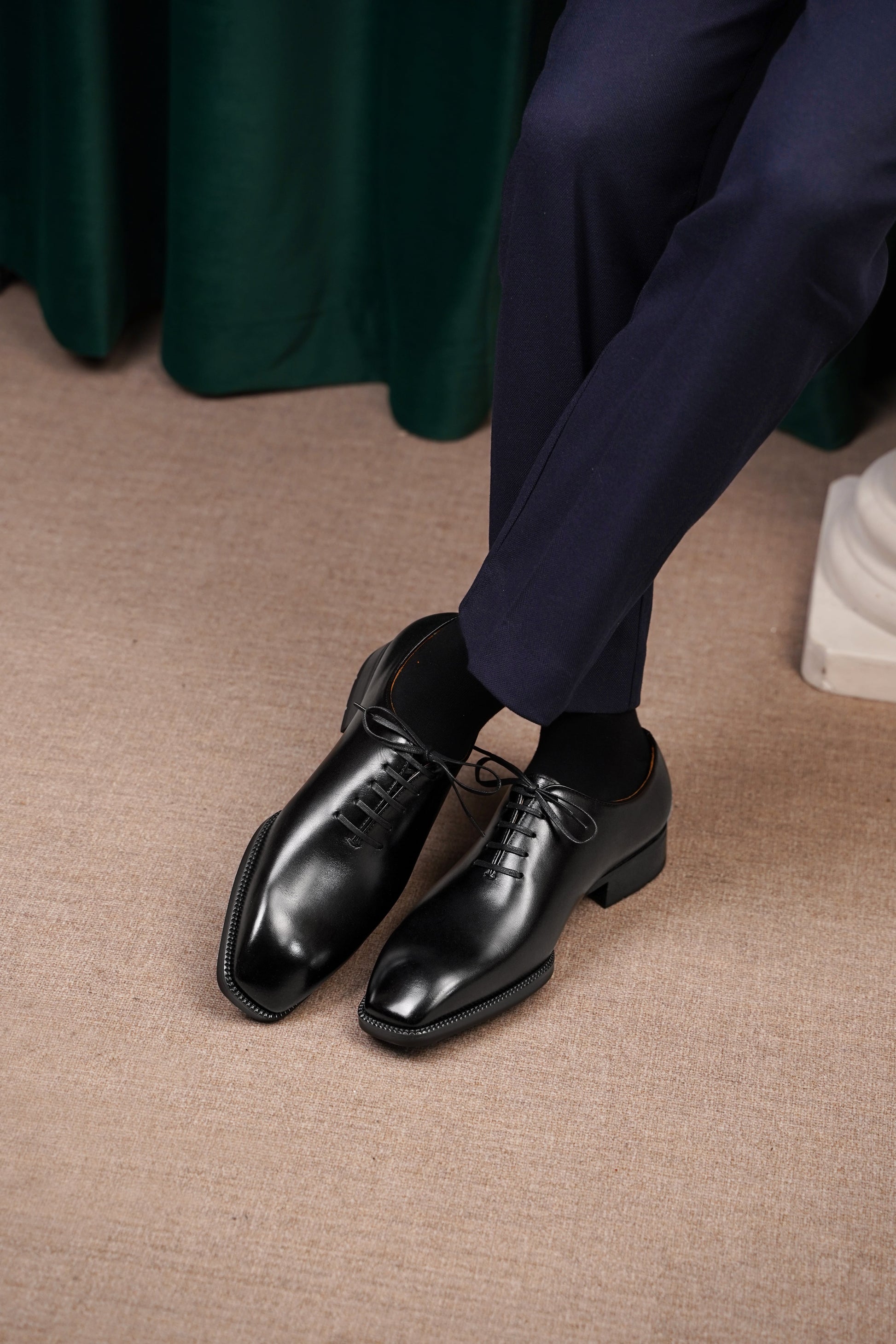 The Classic Black Wholecut Oxford – a view of the shoes being worn by a person in dark blue formal trousers, sitting on a light-colored carpet.