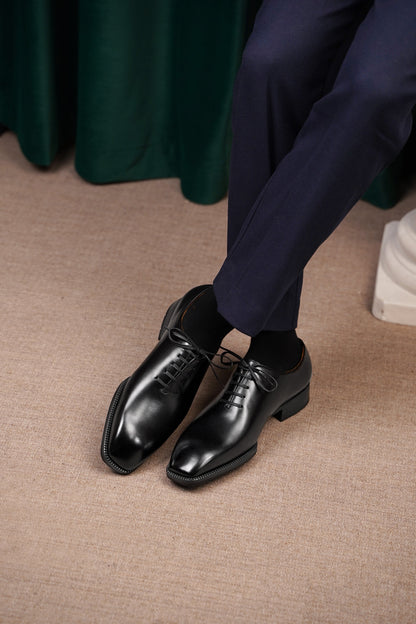 The Classic Black Wholecut Oxford – a view of the shoes being worn by a person in dark blue formal trousers, sitting on a light-colored carpet.