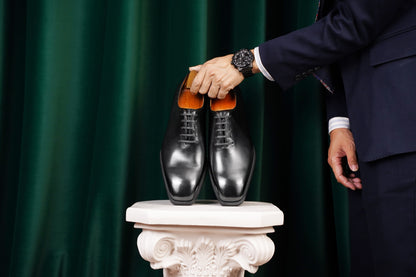 The Classic Black Wholecut Oxford – a person in a navy blue suit holding the pair of wholecut oxfords on a white pedestal, against a dark green backdrop.