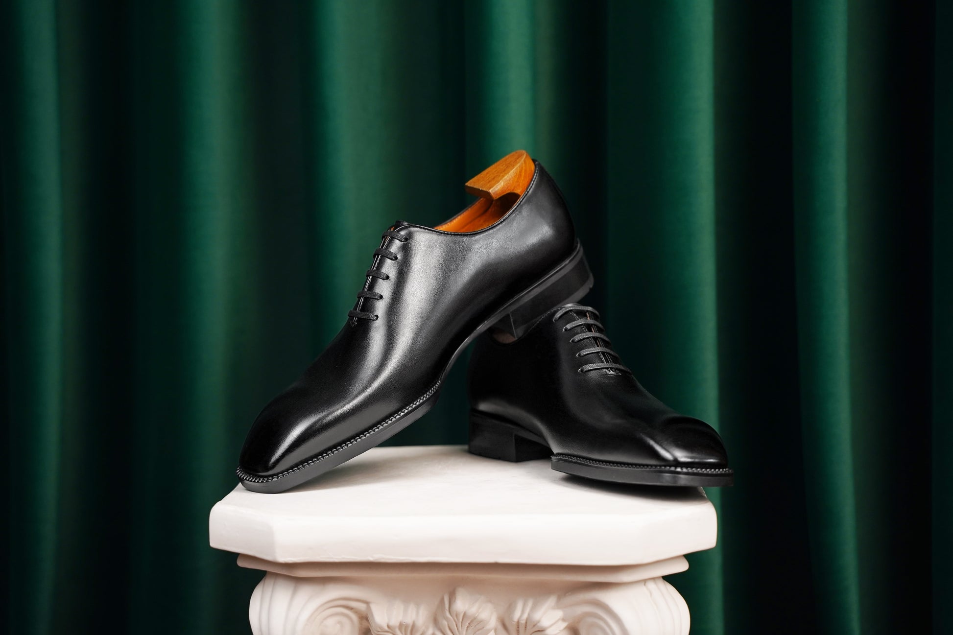 The Classic Black Wholecut Oxford – a side-profile shot of a pair of elegant footwear on a white classical pedestal against a dark green velvet curtain backdrop.