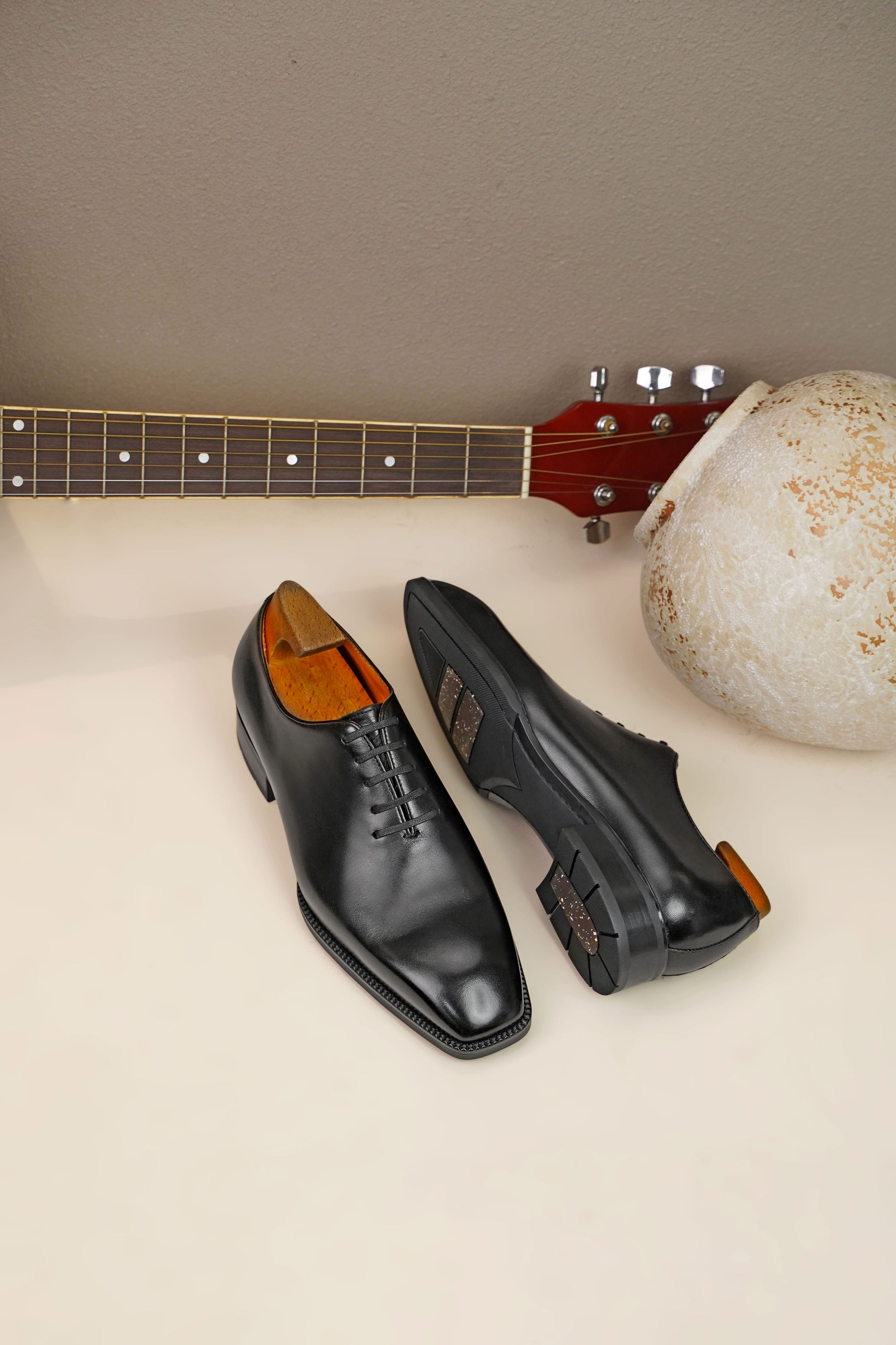 The Classic Black Wholecut Oxford – a side view of a pair of shoes on a light surface, with one shoe turned over to display the sole and a decorative vase in the background.