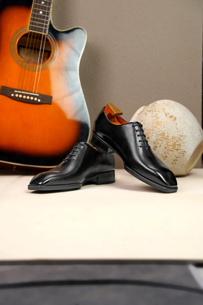 The Classic Black Wholecut Oxford – a dynamic arrangement of the pair of shoes on a light surface, with an acoustic guitar and an artistic vase in the background.