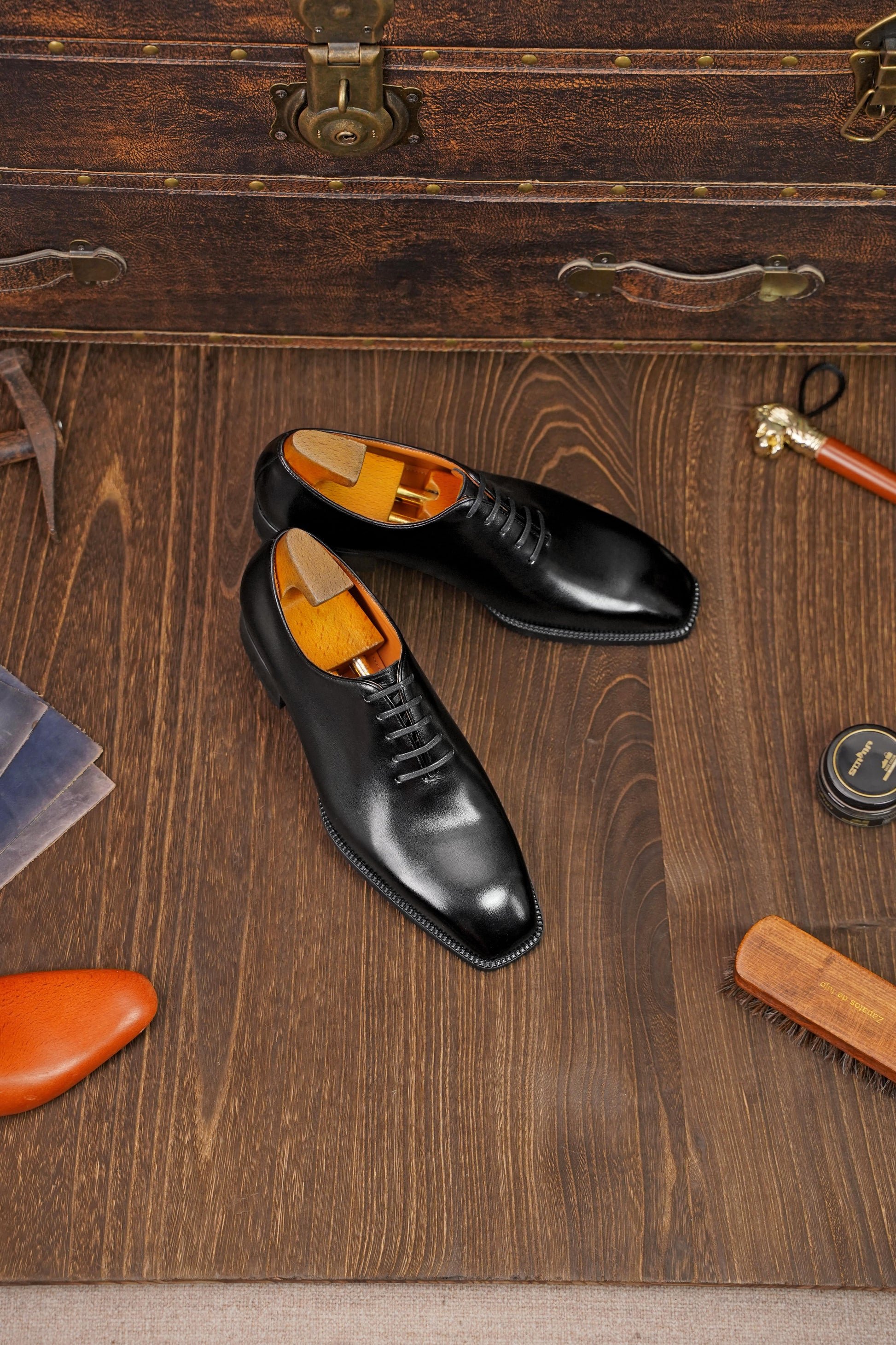 Classic Black Leather Wholecut Oxford Dress Shoes – an angled view of the pair of shoes on a wooden surface, surrounded by shoe care accessories and a vintage suitcase.