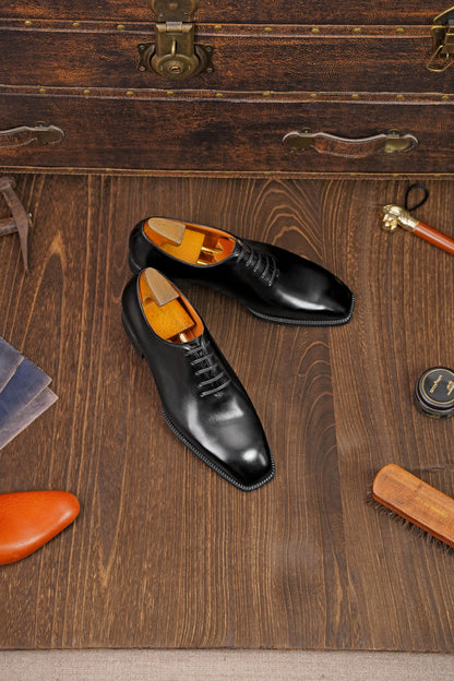 Classic Black Leather Wholecut Oxford Dress Shoes – an angled view of the pair of shoes on a wooden surface, surrounded by shoe care accessories and a vintage suitcase.