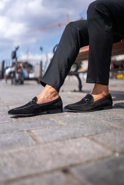 Classic Black Suede Bit Loafers for Office & Evening Wear – worn by a man in black trousers, sitting on a bench on a stone-paved ground, highlighting a classic loafer design and metal bit.