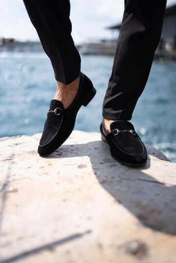 Classic Black Suede Bit Loafers for Office & Evening Wear – worn by a man in black trousers, standing on a ledge by the water, showcasing timeless elegance and metal bit detail.
