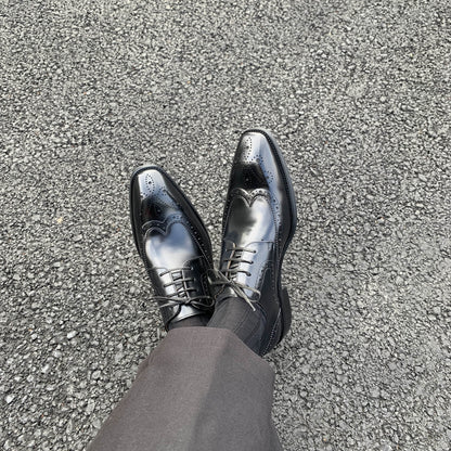 Men's Classic Black Wingtip Brogue Derby Shoes – worn with black trousers on asphalt, showcasing the elegant wingtip design from above.