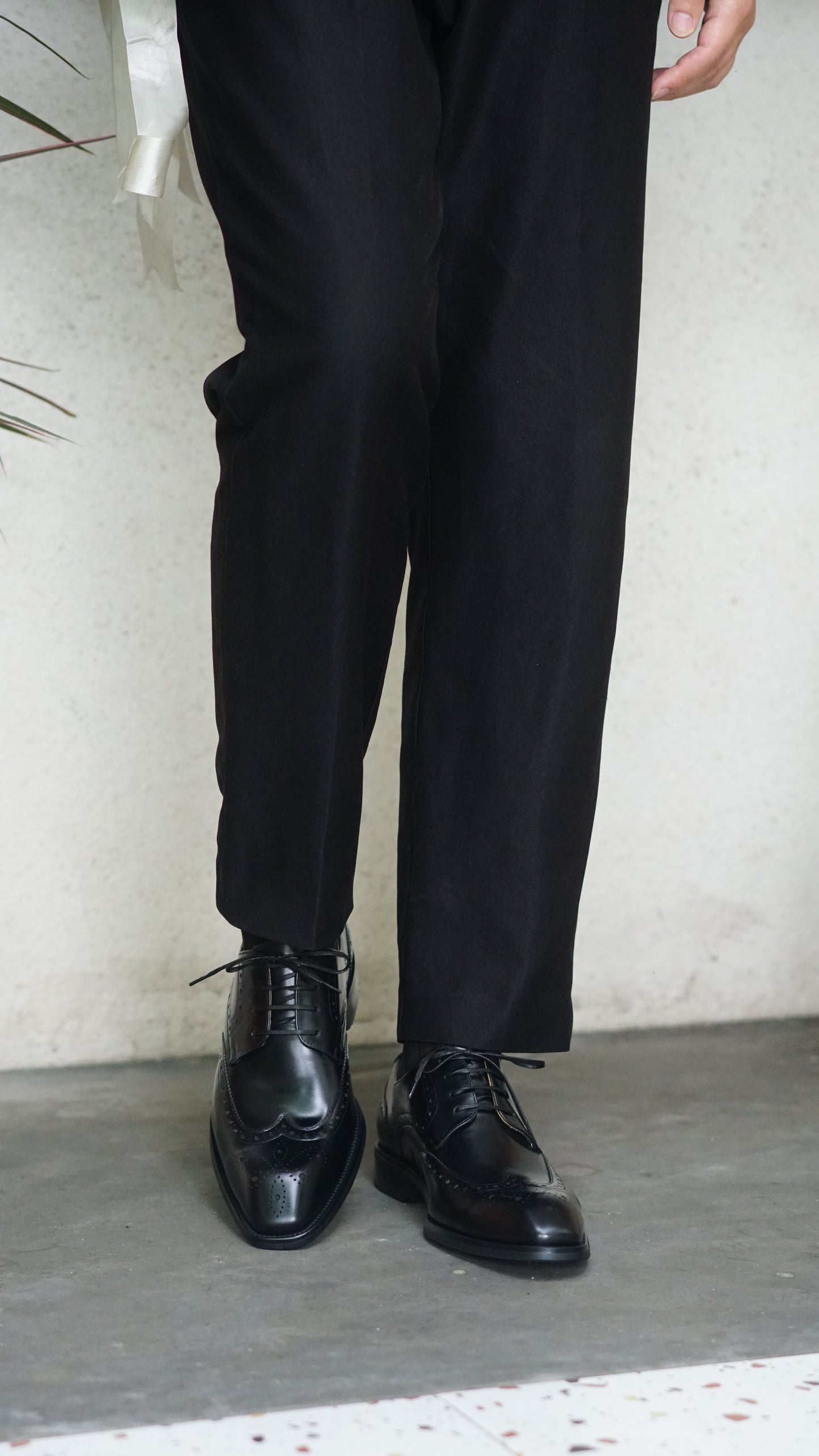 Men's Classic Black Wingtip Brogue Derby Shoes – sleek black leather, worn with black trousers, standing on a light-colored surface.