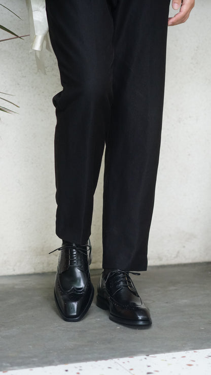 Men's Classic Black Wingtip Brogue Derby Shoes – sleek black leather, worn with black trousers, standing on a light-colored surface.