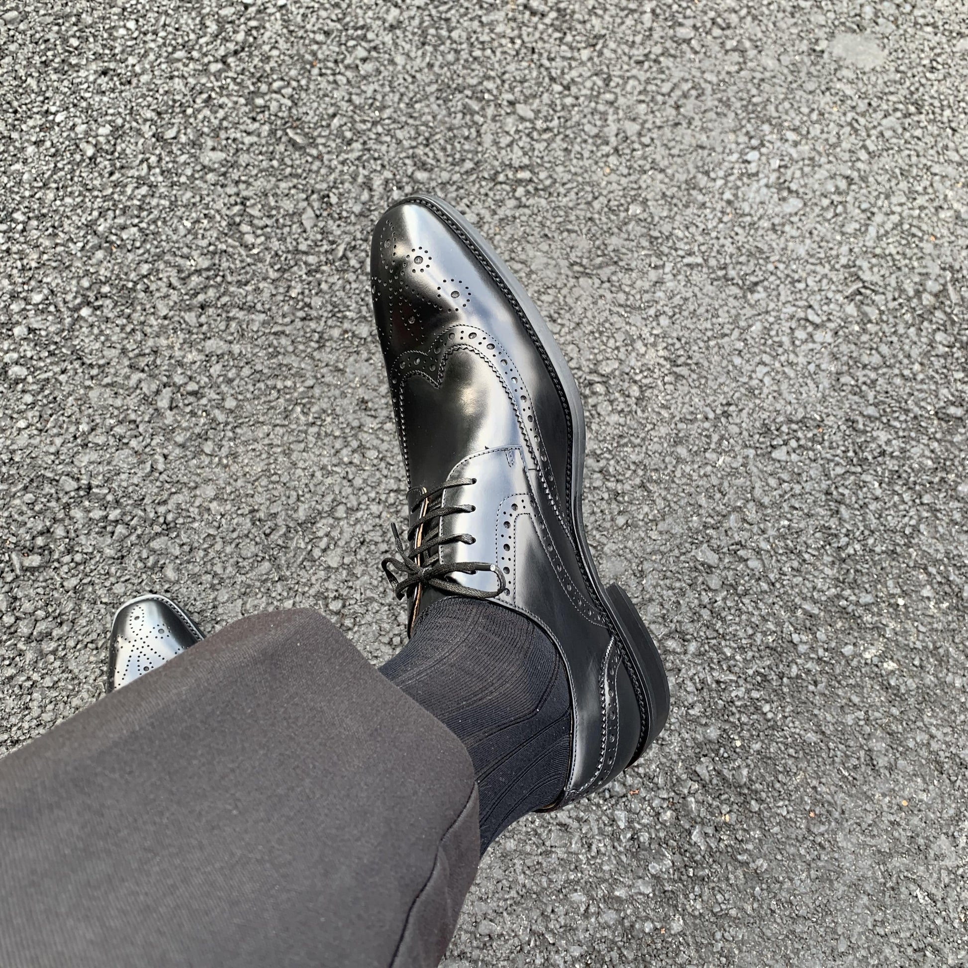 Men's Classic Black Wingtip Brogue Derby Shoes – worn with black trousers on asphalt, showcasing the elegant wingtip design from above.
