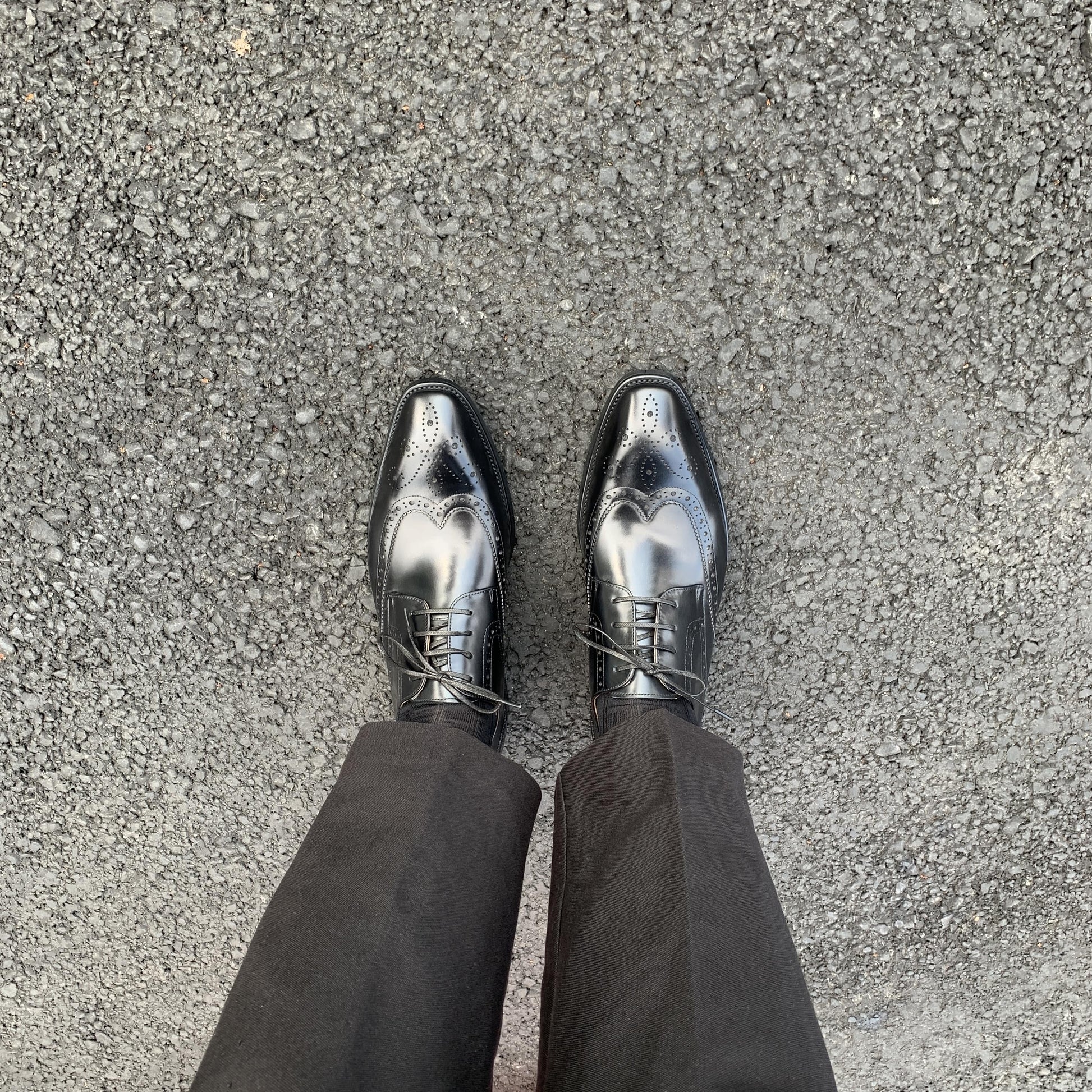 Men's Classic Black Wingtip Brogue Derby Shoes – overhead view on asphalt, showcasing the sleek silhouette and wingtip design on a man wearing black trousers.
