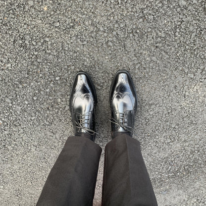 Men's Classic Black Wingtip Brogue Derby Shoes – overhead view on asphalt, showcasing the sleek silhouette and wingtip design on a man wearing black trousers.