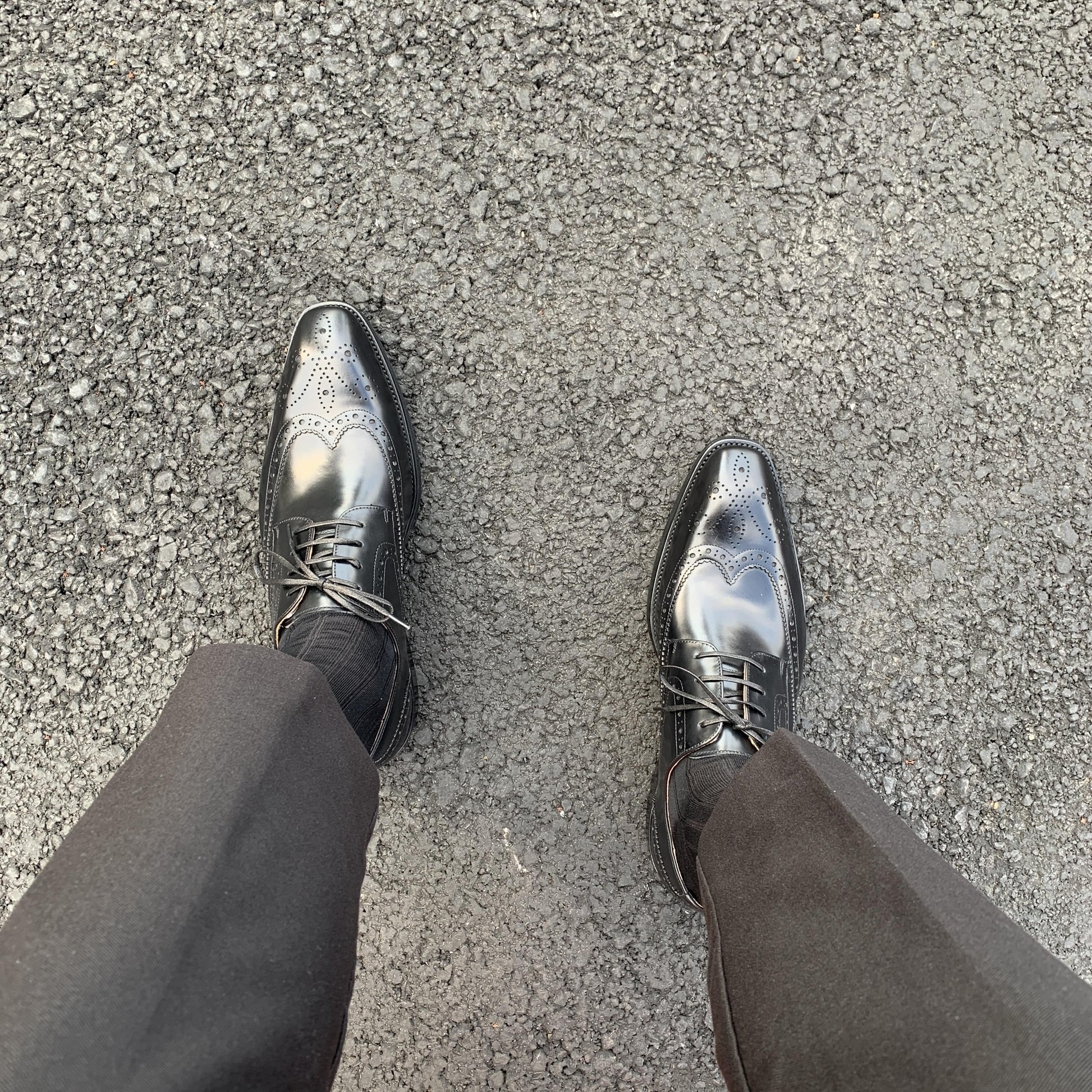Men's Classic Black Wingtip Brogue Derby Shoes – top-down view on asphalt, highlighting polished finish and intricate broguing on a man wearing black trousers.