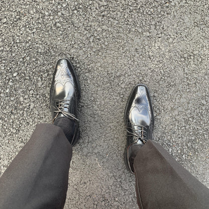 Men's Classic Black Wingtip Brogue Derby Shoes – top-down view on asphalt, highlighting polished finish and intricate broguing on a man wearing black trousers.