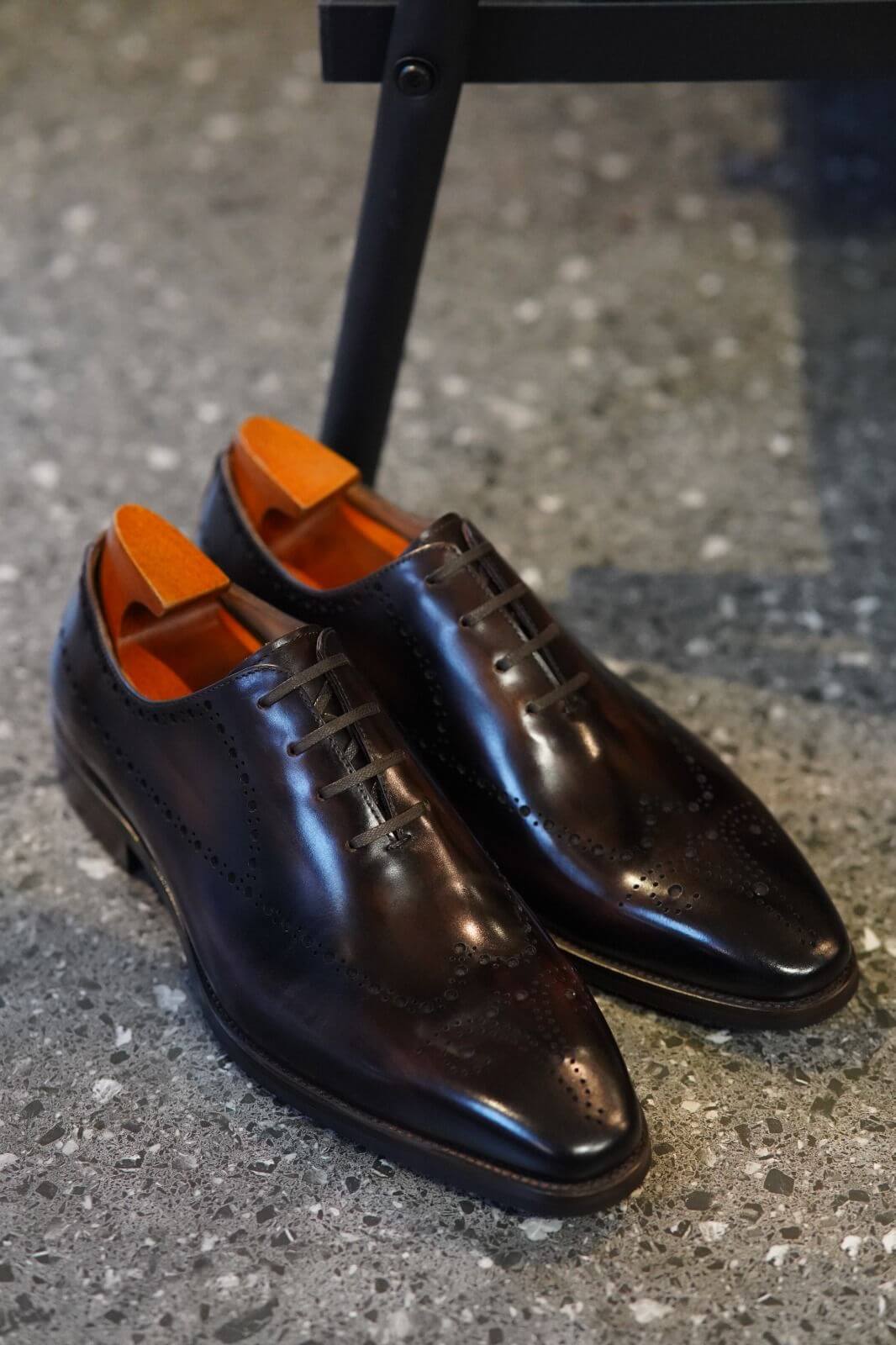 Pair of polished black dress shoes with orange soles on a textured surface