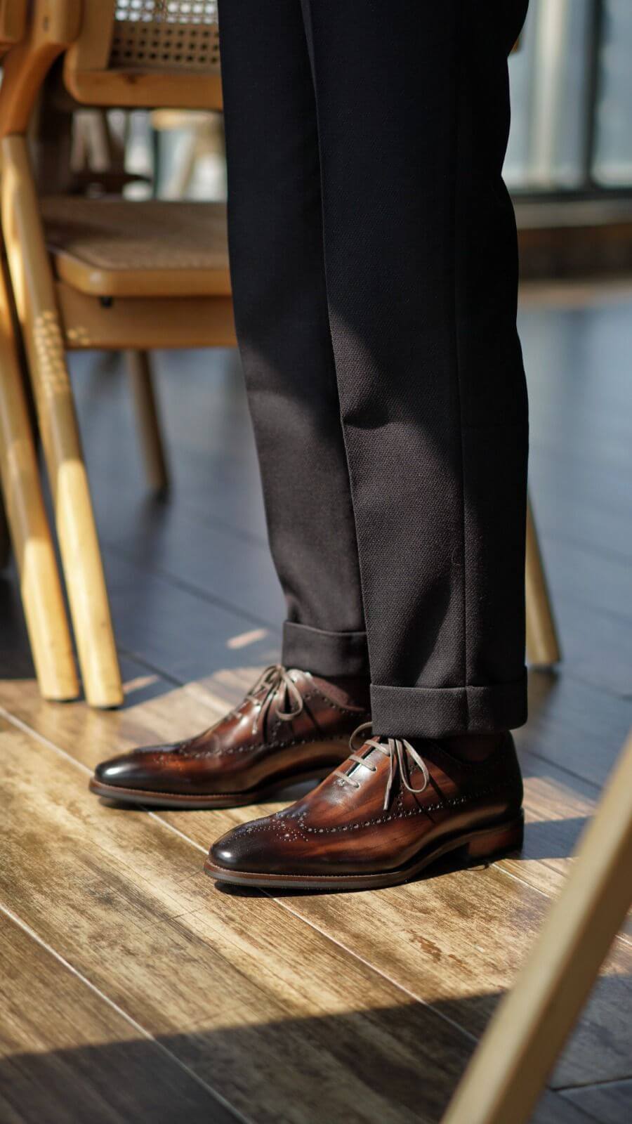Brown leather shoes and dark pants on a wooden floor with chairs in the background