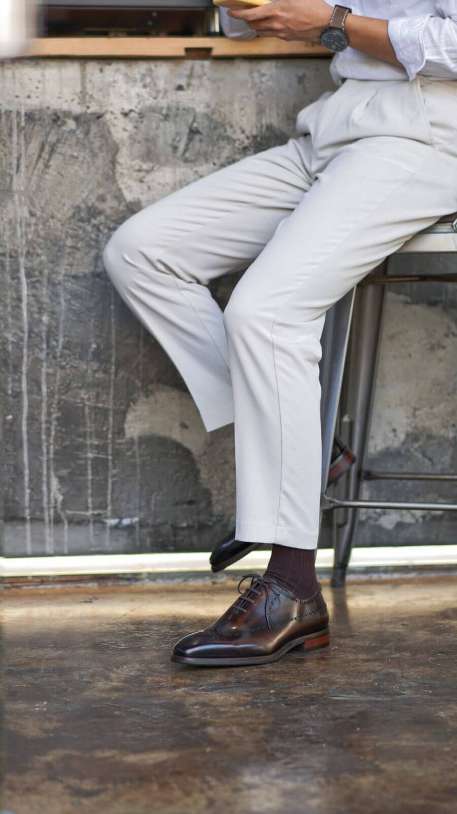 Person sitting on a stool wearing light-colored pants and brown shoes, with a concrete wall in the background.