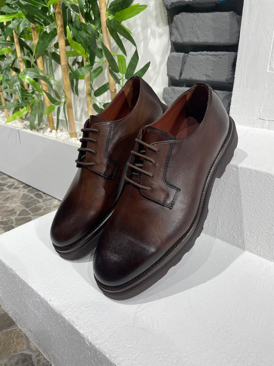 Classic Brown Leather Men's Derby Shoes – sleek design with chunky sole, displayed on a white ledge against a grey brick wall and green bamboo.