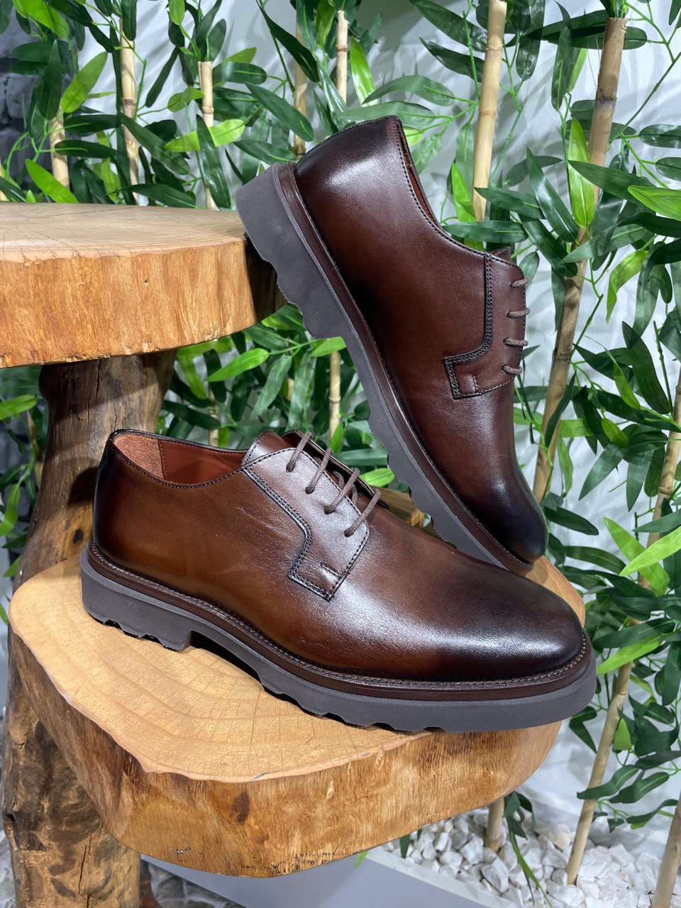 Classic Brown Leather Men's Derby Shoes – premium full-grain leather, displayed on a white ledge with green bamboo and a brick wall background.