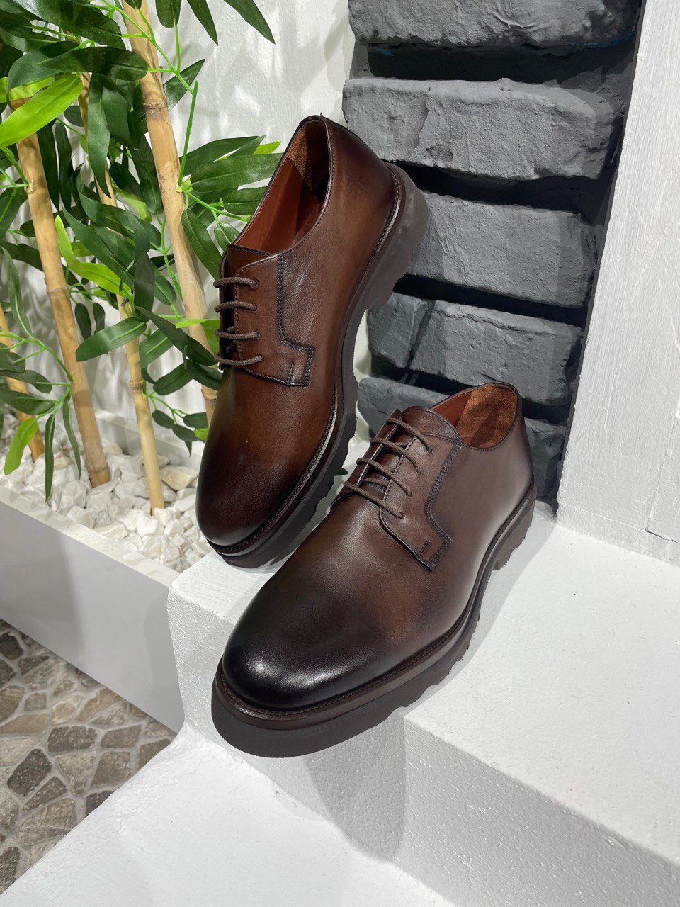 Classic Brown Leather Men's Derby Shoes – rich brown full-grain leather, displayed on a white ledge with a brick wall and green bamboo.