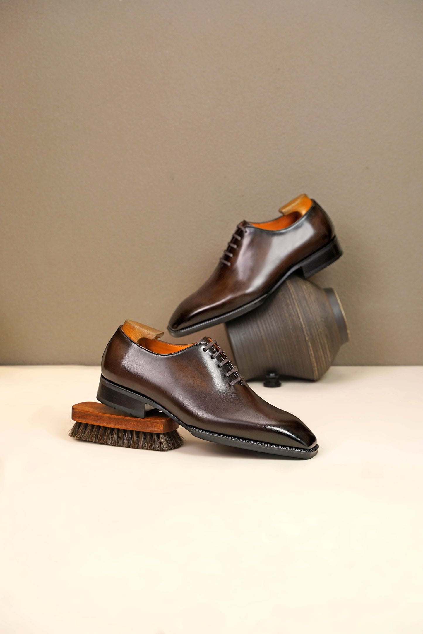 Classic Brown Leather Wholecut Oxford Dress Shoe – a side-profile view of a single shoe standing on a light surface, with an acoustic guitar and an artistic vase in the background.