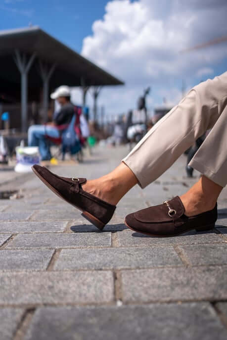 Classic Brown Suede Bit Loafers for Office & Evening Wear – worn by a man in light trousers, sitting on a bench on a stone-paved ground, highlighting a classic loafer design and metal bit.