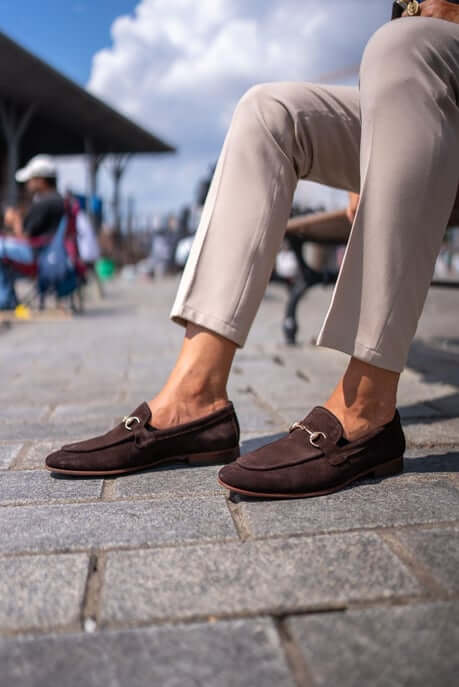 Classic Brown Suede Bit Loafers for Office & Evening Wear – worn by a man in light trousers, standing on a ledge by the water, showcasing timeless elegance and metal bit detail.