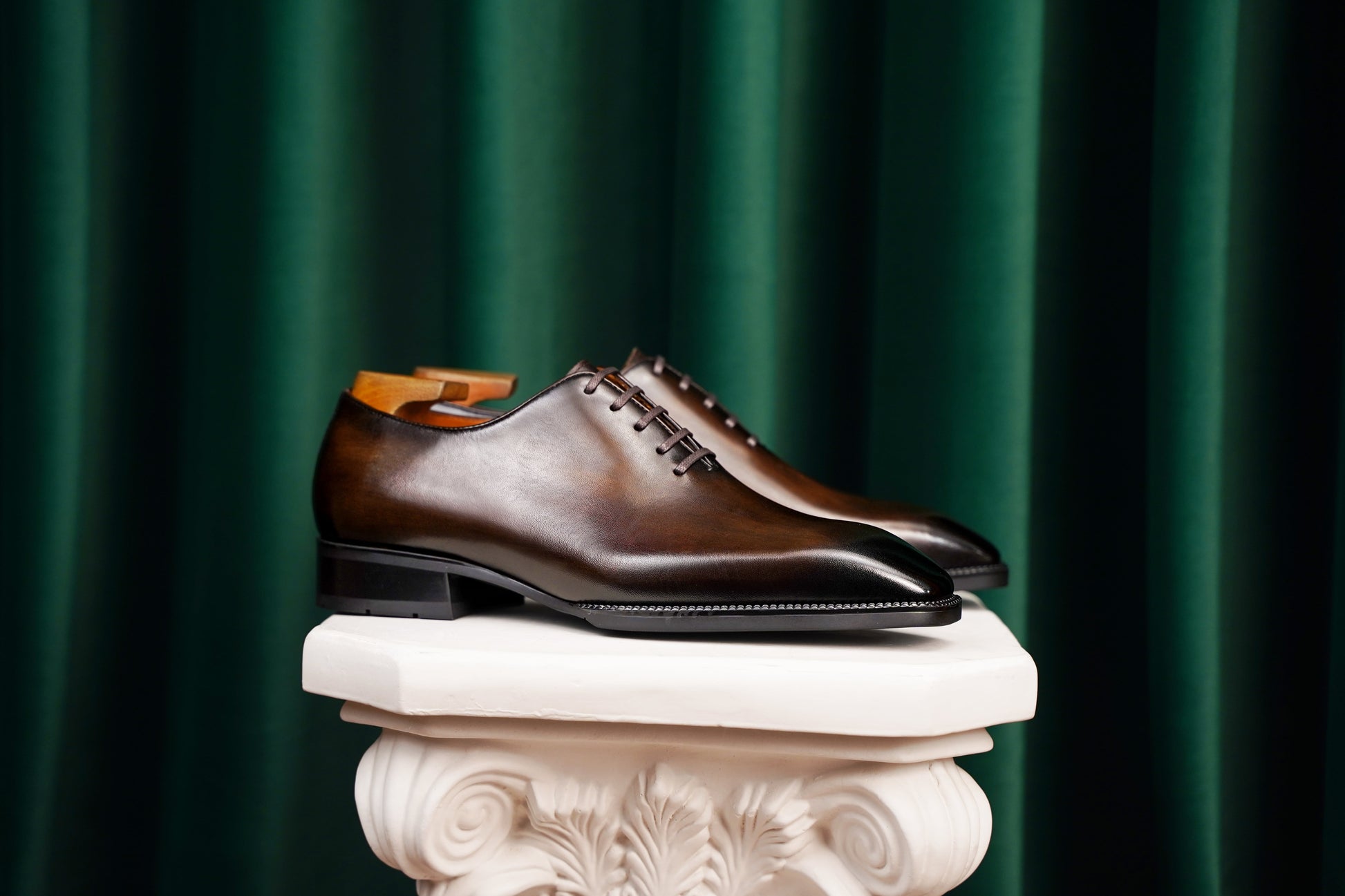 Classic Burnished Brown Leather Wholecut Oxford Dress Shoes – a side-profile shot of a pair of elegant footwear on a white classical pedestal against a dark green velvet curtain backdrop.