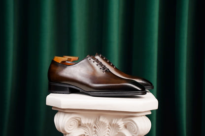 Classic Burnished Brown Leather Wholecut Oxford Dress Shoes – a side-profile shot of a pair of elegant footwear on a white classical pedestal against a dark green velvet curtain backdrop.
