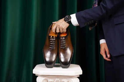 Classic Burnished Brown Leather Wholecut Oxford Dress Shoes – a person in a navy blue suit holding the pair of wholecut oxfords on a white pedestal, against a dark green backdrop.