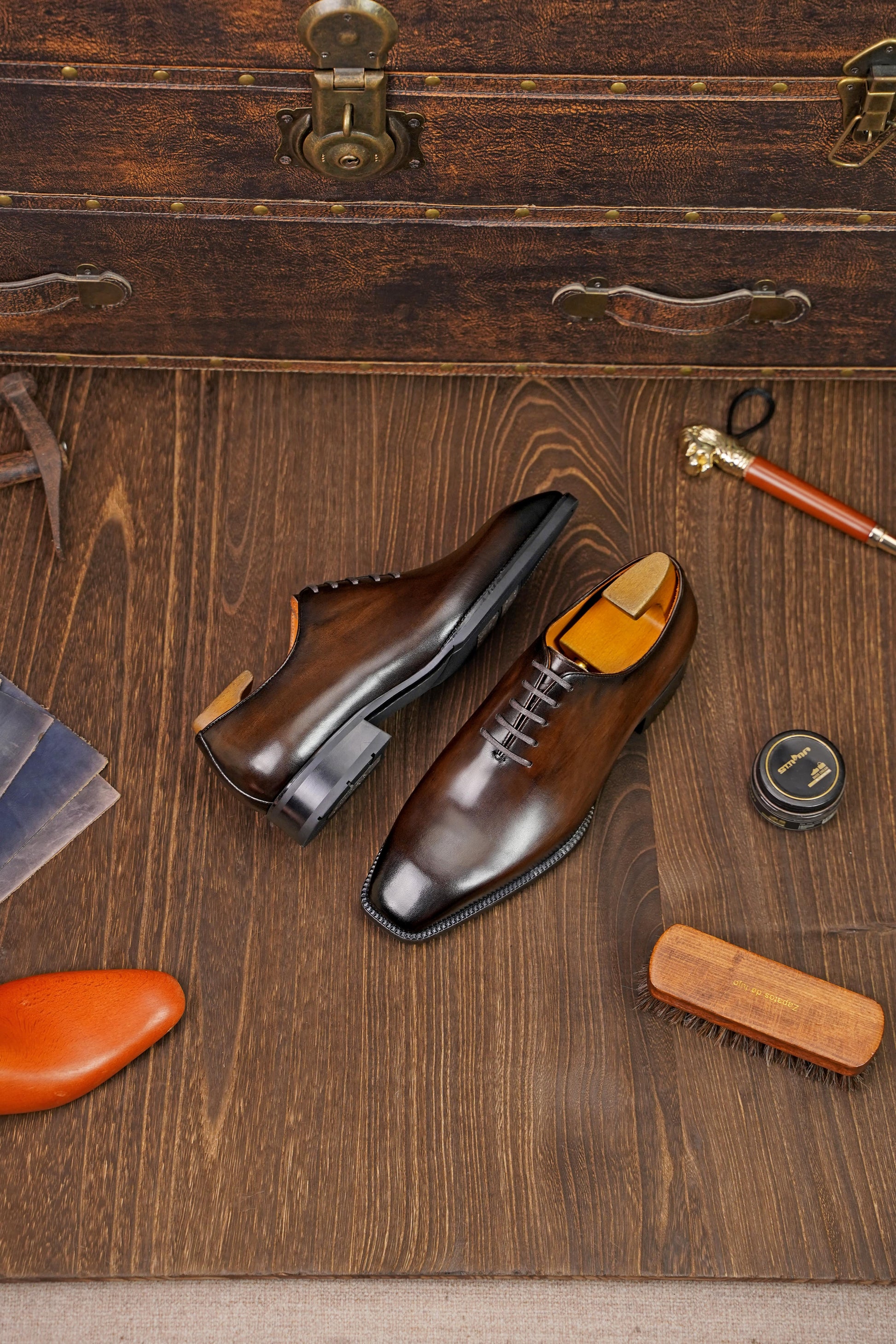 Classic Burnished Brown Leather Wholecut Oxford Dress Shoes – an angled view of the pair of shoes on a wooden surface, surrounded by shoe care accessories and a vintage suitcase.