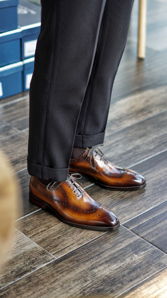 Brown dress shoes worn with dark trousers on a wooden floor.