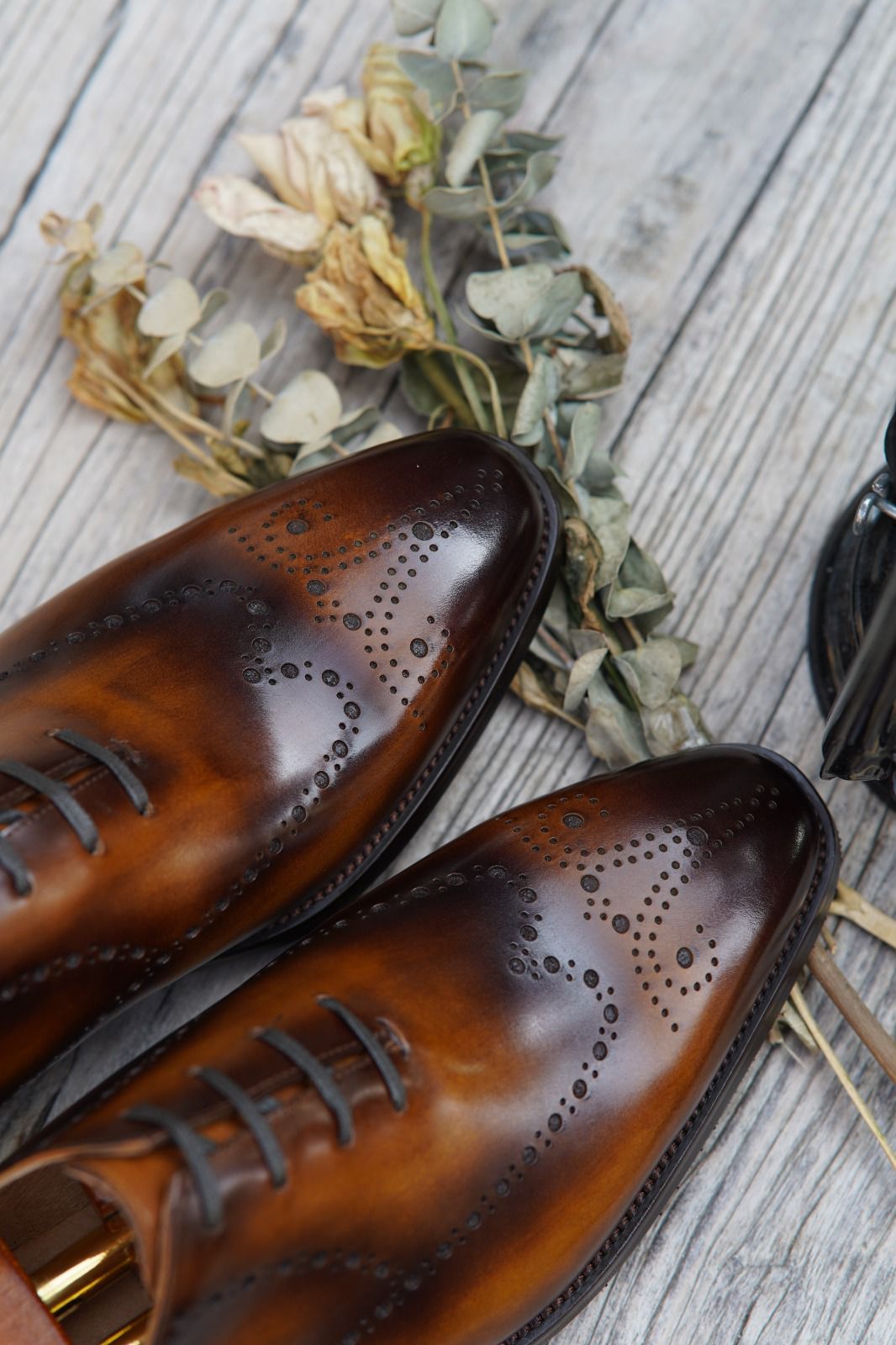 Brown leather shoes with gold accents on a wooden surface with dried flowers.