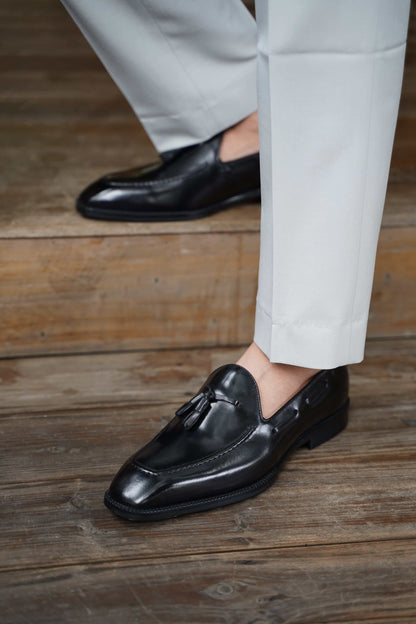 Elegant Hand-Made Black Leather Tassel Loafers – close-up of a man's feet in polished black leather shoes, worn with light cuffed trousers on a wooden floor.