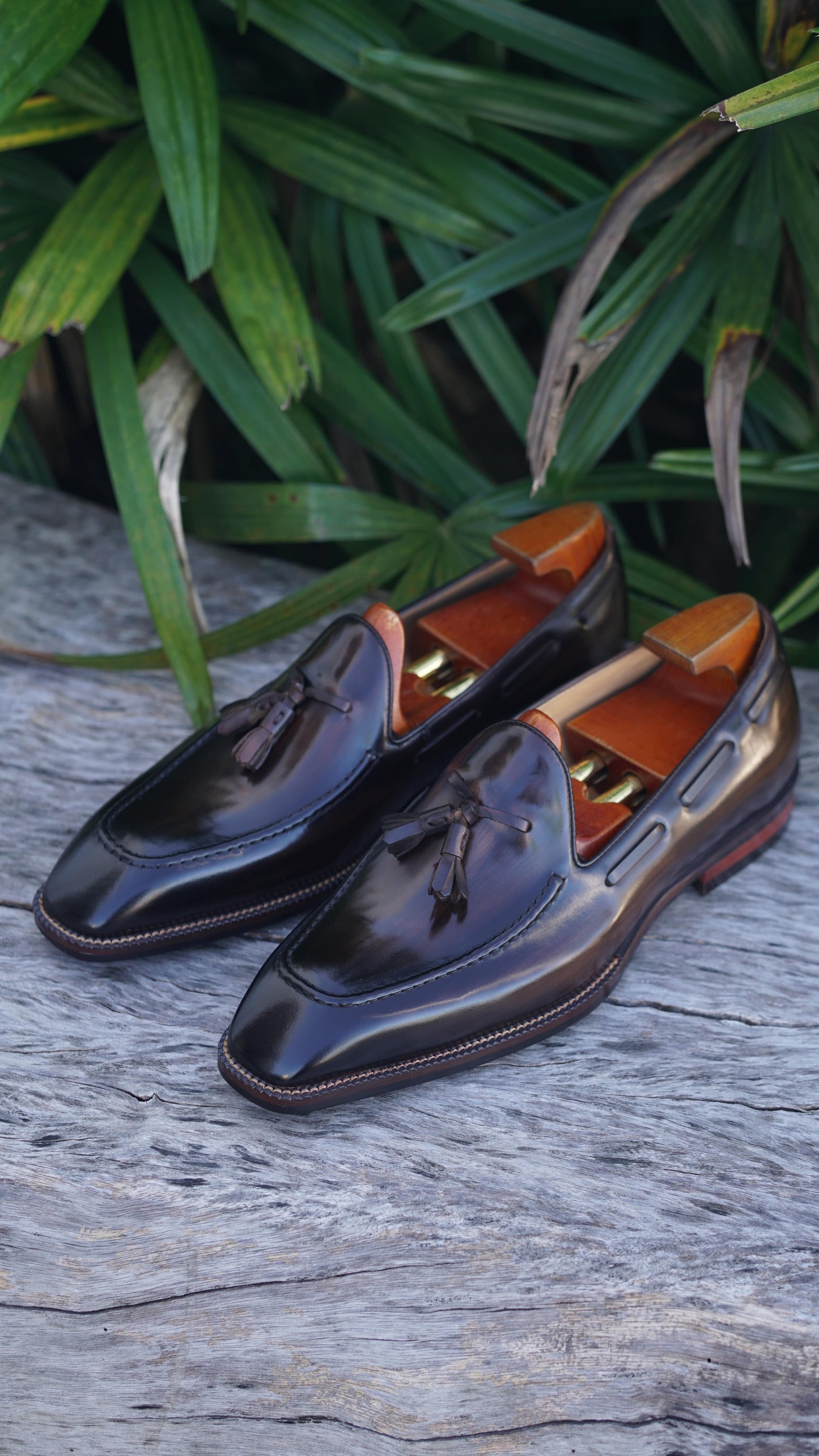 Hand-Made Dark Brown Leather Tassel Loafers – pair with shoe trees on a wooden surface, with green foliage, highlighting sophisticated design.